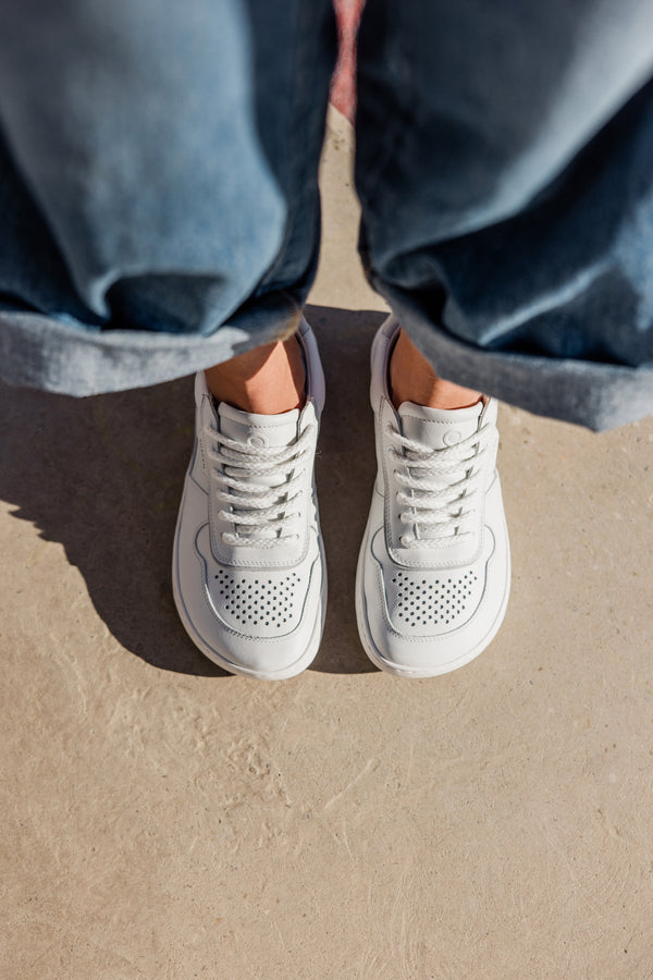 A top-down view of a person wearing white low-top sneakers with blue jeans, standing on a light concrete surface.