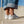 A person wearing light blue cuffed jeans and white low-top sneakers, standing on a concrete ledge outdoors against a blue sky.