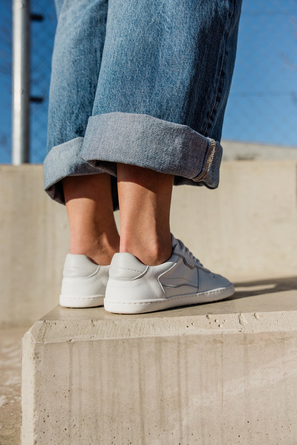 A person wearing light blue cuffed jeans and white low-top sneakers, standing on a concrete ledge outdoors against a blue sky.