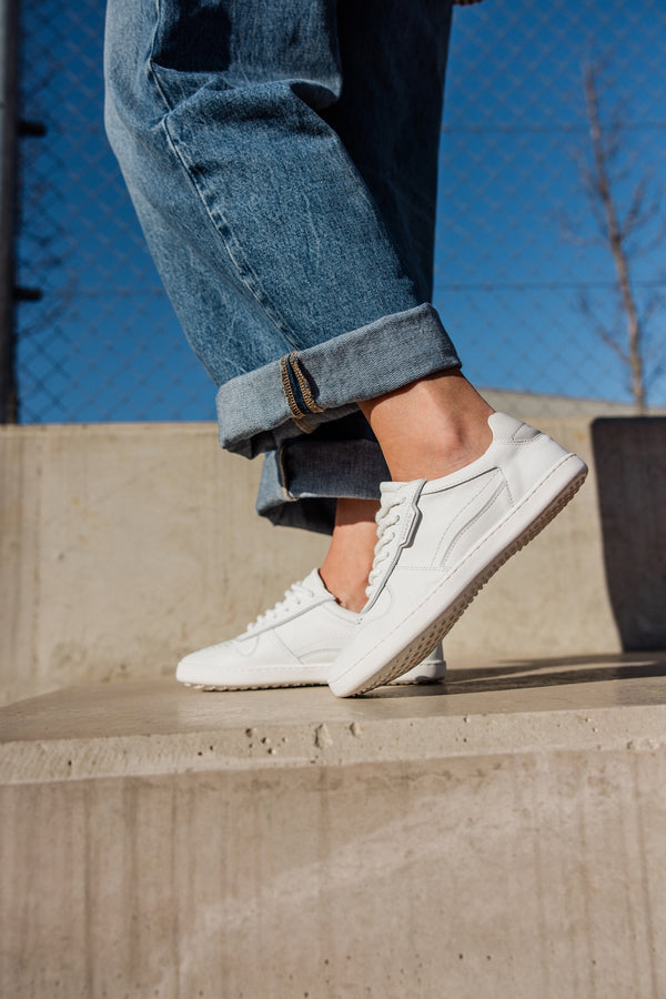 White leather low-top sneakers worn with cuffed blue jeans, shown outdoors on concrete steps under a clear blue sky.